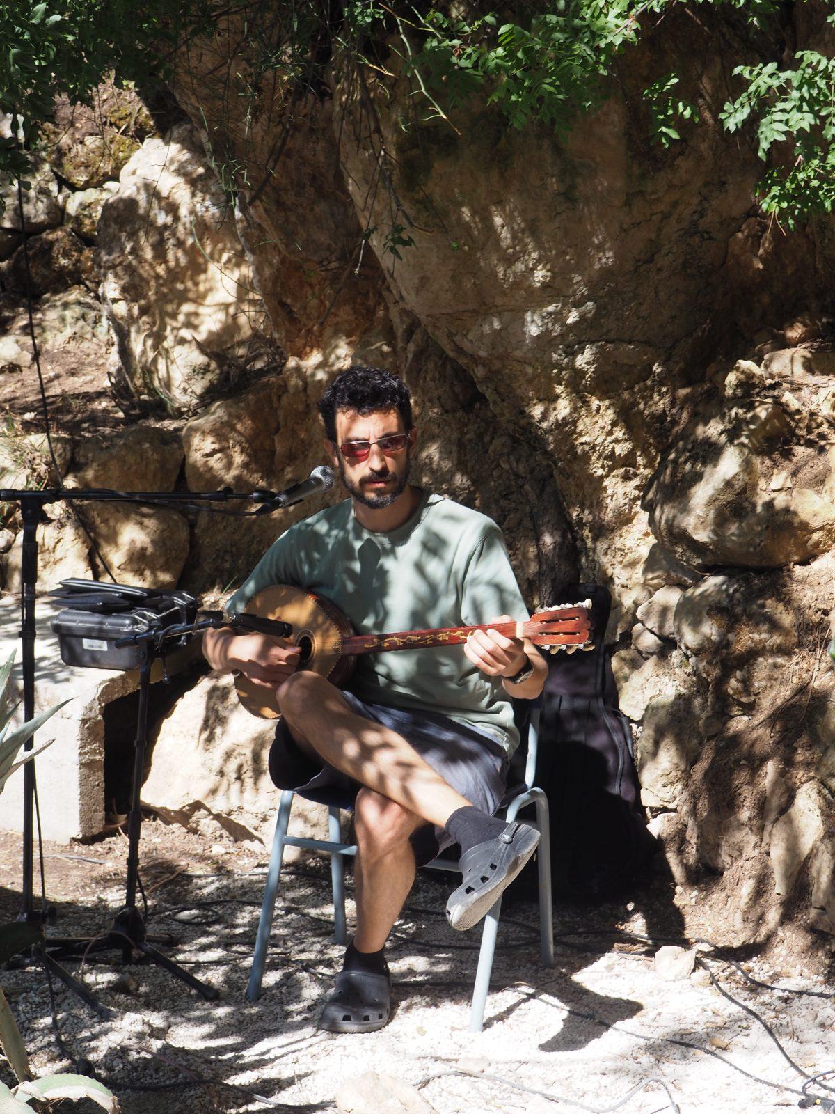 Um homem de t-shirt e calções, sentado numa cadeira e tocando um instrumento de cordas de origem local. 