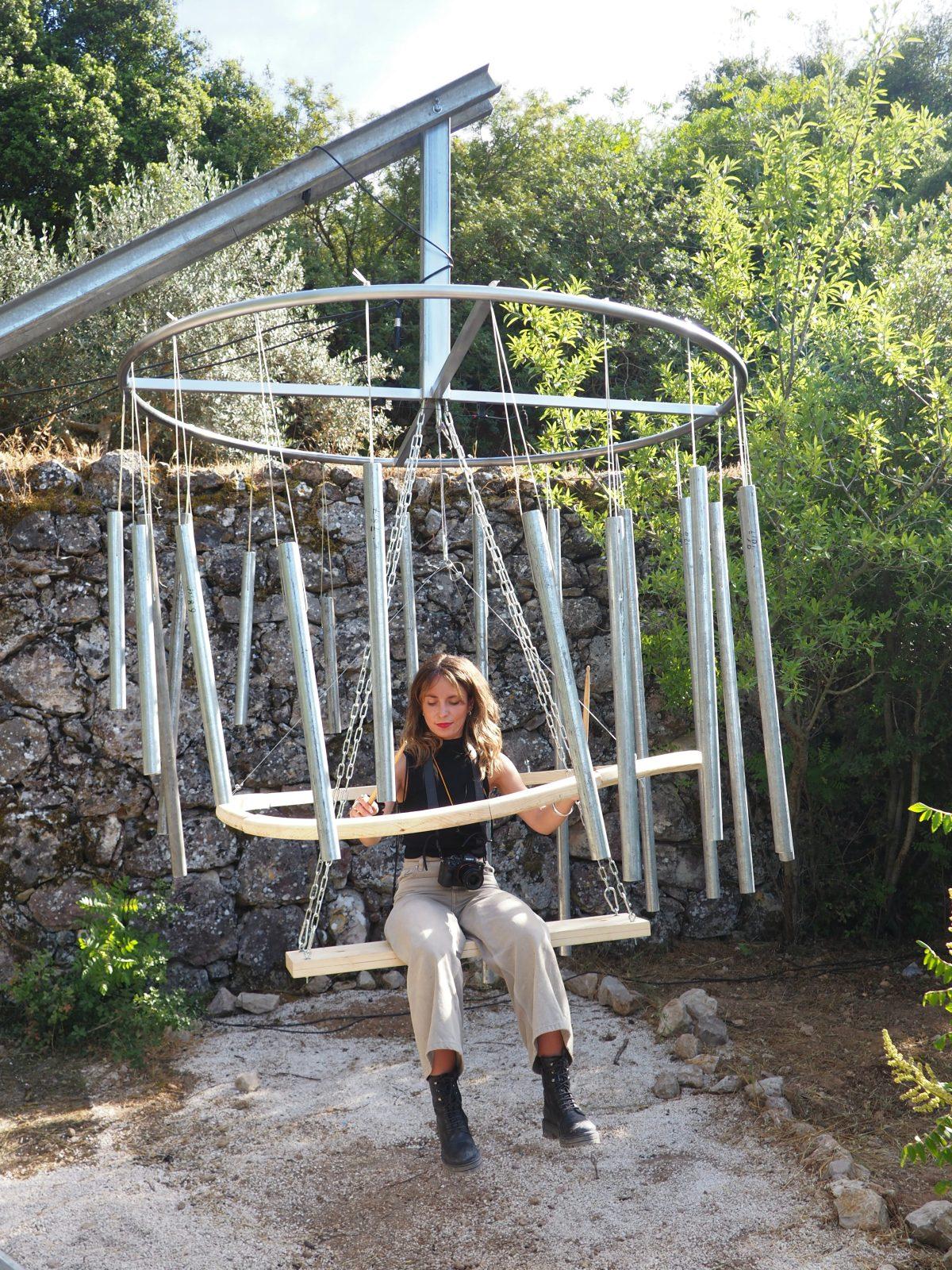 Nesta fotografia vemos uma mulher sentada numa espécie de baloiço. Em seu redor estão várias peças metálicas dispostas em círculo. Este baloiço, em conjunto com as peças metálicas, compõem um instrumento de percursão que está a ser tocado pela mulher no baloiço. 