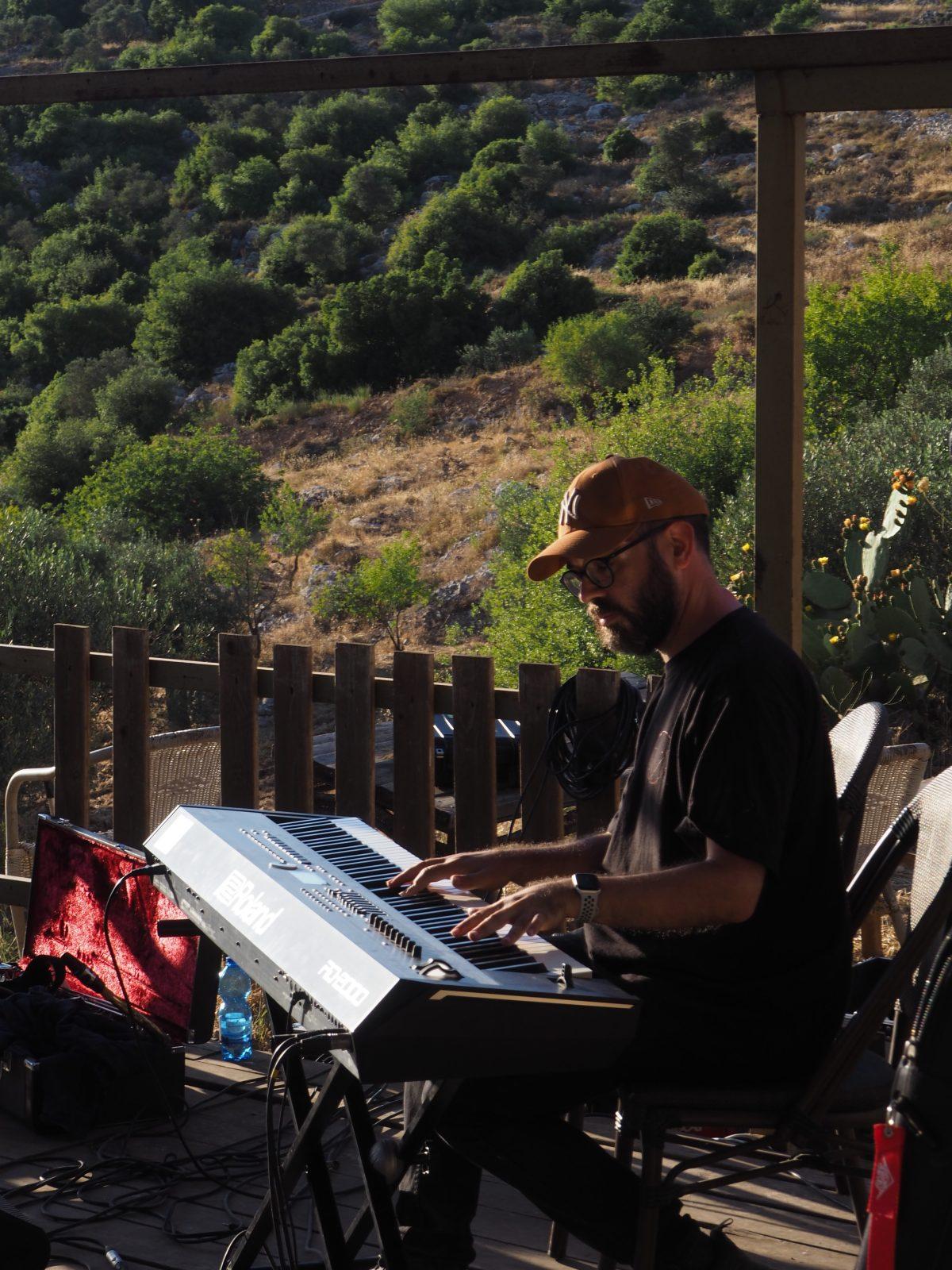 Um homem sentado em frente a um teclado musical, tendo como pano de fundo uma paisagem semelhante aos vales das restantes fotografias. 