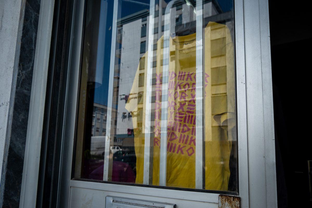 Fotografia da porta do Tabanka, local onde decorreu a entrevista. Nesta fotografia vemos o vidro da porta, onde se pode ver o reflexo dos prédios habitacionais do outro lado da estrada, mas vemos também uma tshirt de merchandising.