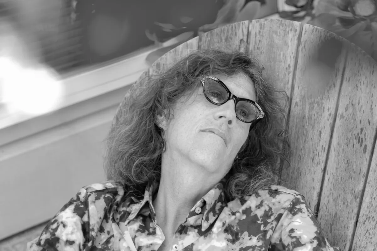 Fotografia a preto e branco onde está a historiadora Susan Stryker, uma mulher branca de meia idade, sentada numa cadeira de jardim de madeira. Susan encontra-se reclinada e veste uma camisa com motivos florais, tem o cabelo solto pelos ombros e uns óculos cat-eye pretos com brilhantes nas pontas.